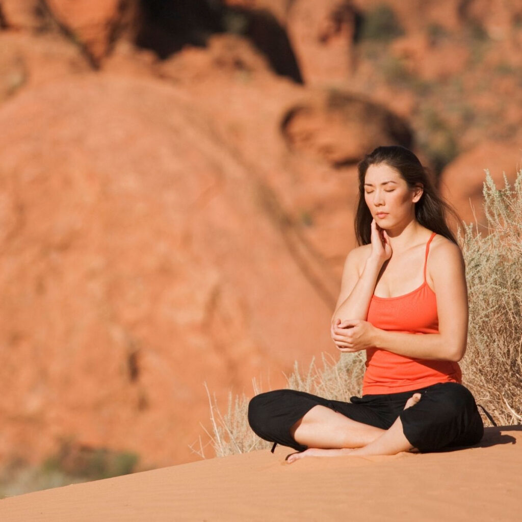 A dark haired woman in an orange tank top meditating in the desert.