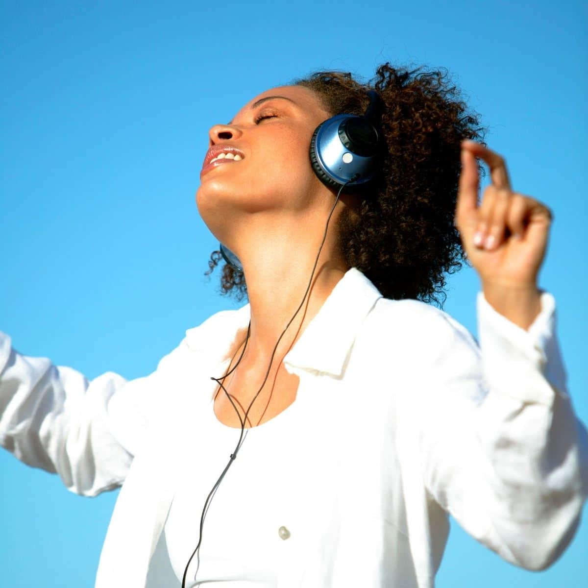 A black woman wearing a white shirt dancing with headphones in front of a blue background.