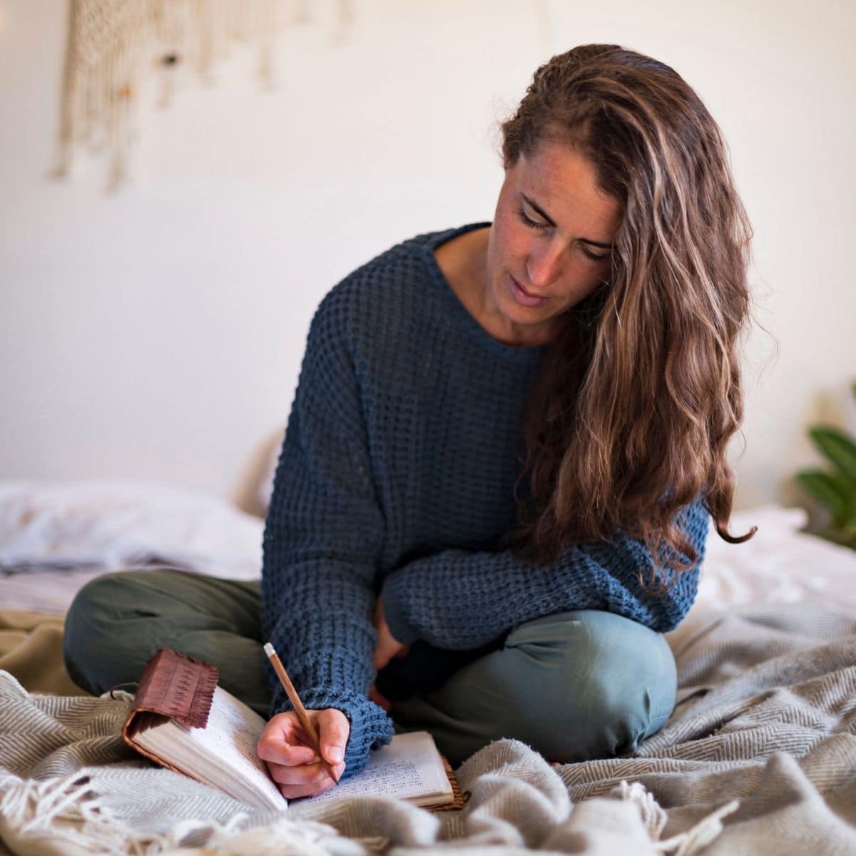 A woman with long brown hair writing in a journal while sitting on the bed.