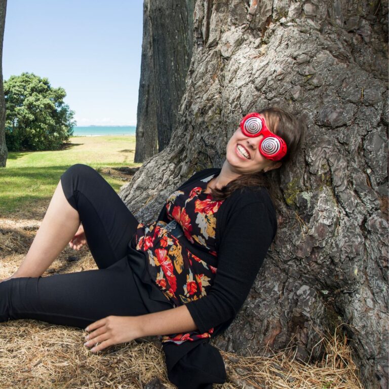 A brown haired woman leaning against a tree wearing crazy spiral glasses.